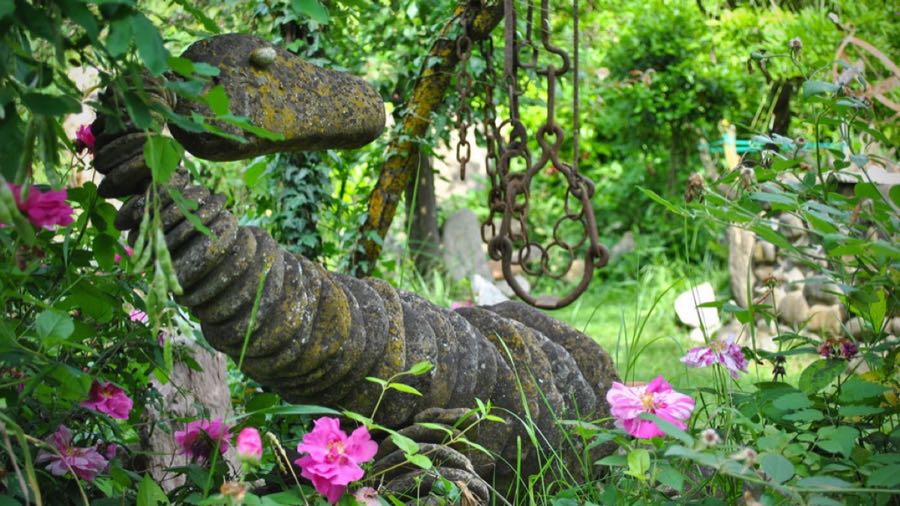 Scultura dal lungo dorso a dischi impilati, con testa allungata, tra i fiori del giardino